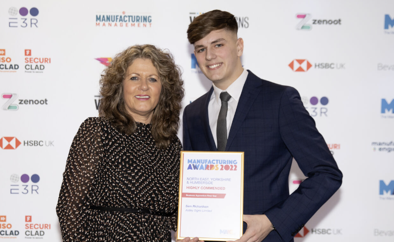 Two people smiling, one holding a framed award, standing in front of a branded backdrop.