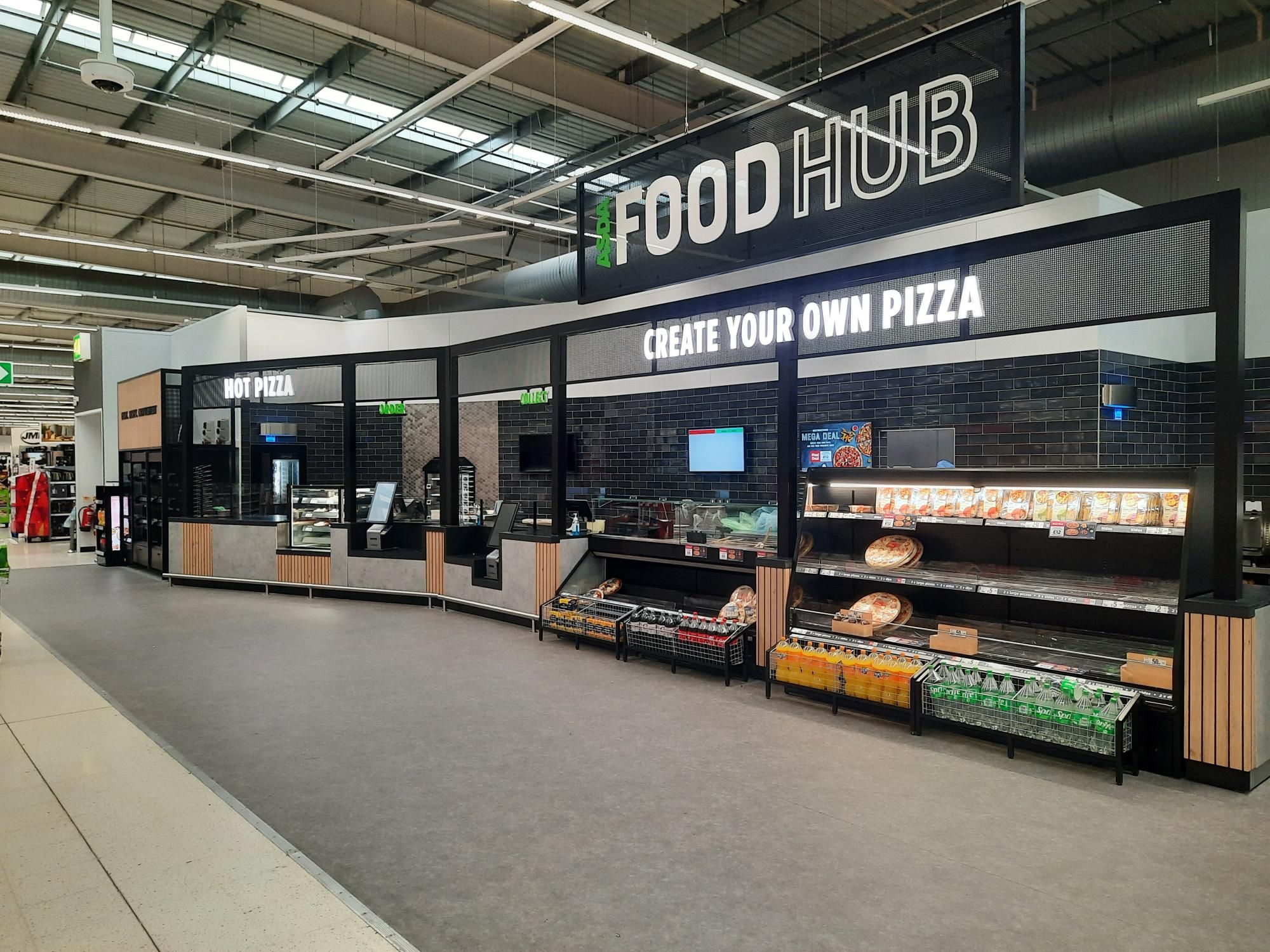 Modern supermarket food hub with "Hot Pizza" and "Create Your Own Pizza" stations. Features digital displays, packaged snacks, pizzas, and fresh vegetables. Bright overhead signage with "Food Hub" branding. Industrial-style black and wood decor.