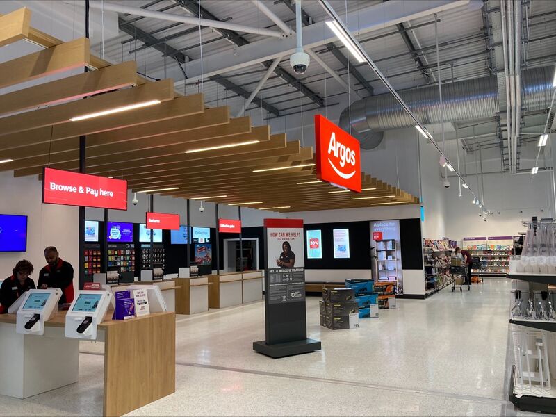 Interior of an Argos store with digital kiosks and red signage under wooden ceiling panels.