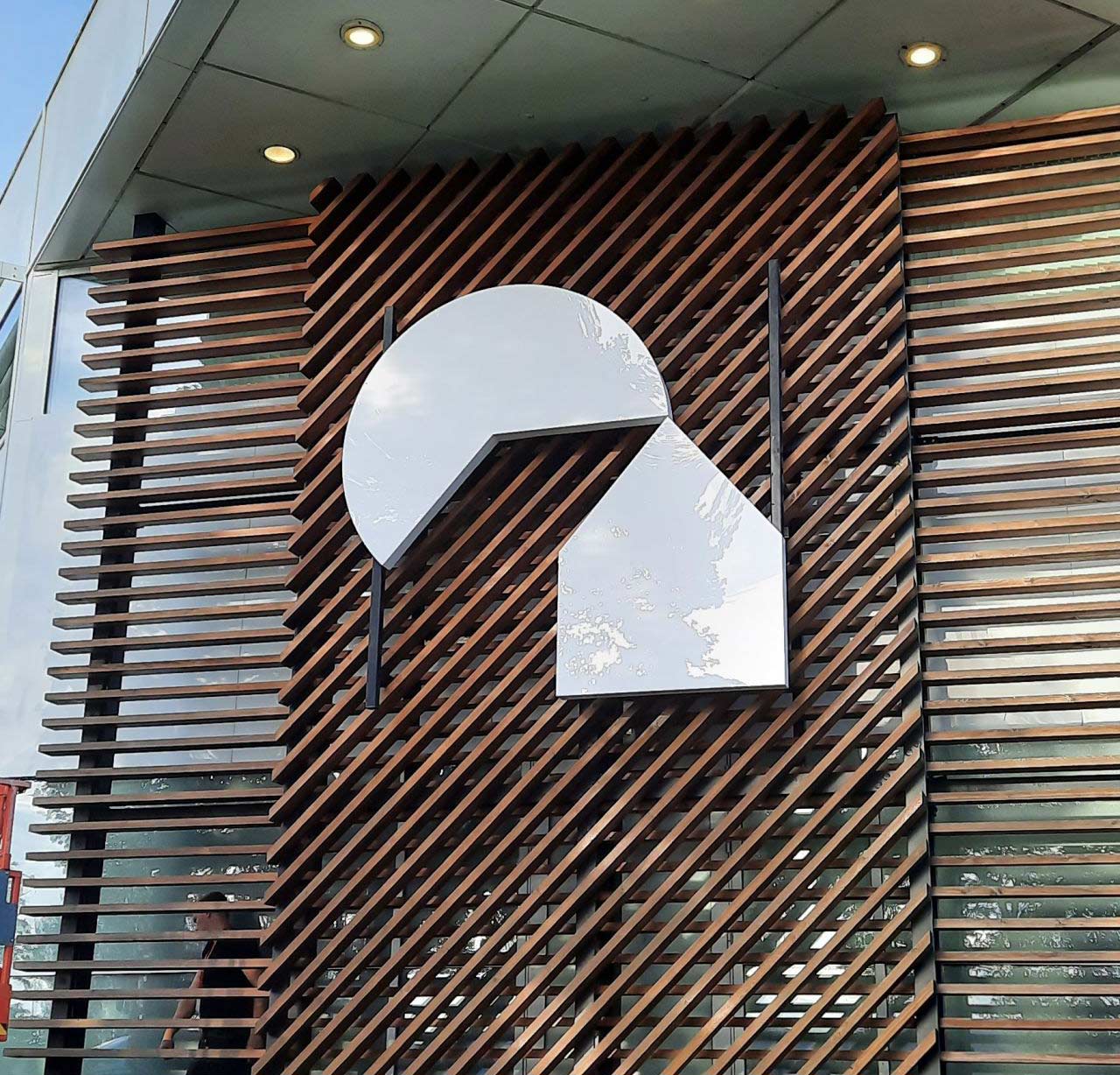 Exterior view of a modern building facade featuring vertical and diagonal wooden slats with an abstract geometric logo in white, consisting of a semi-circle and a tilted rhombus. The design is illuminated by recessed ceiling lights, enhancing its contemporary architectural style.