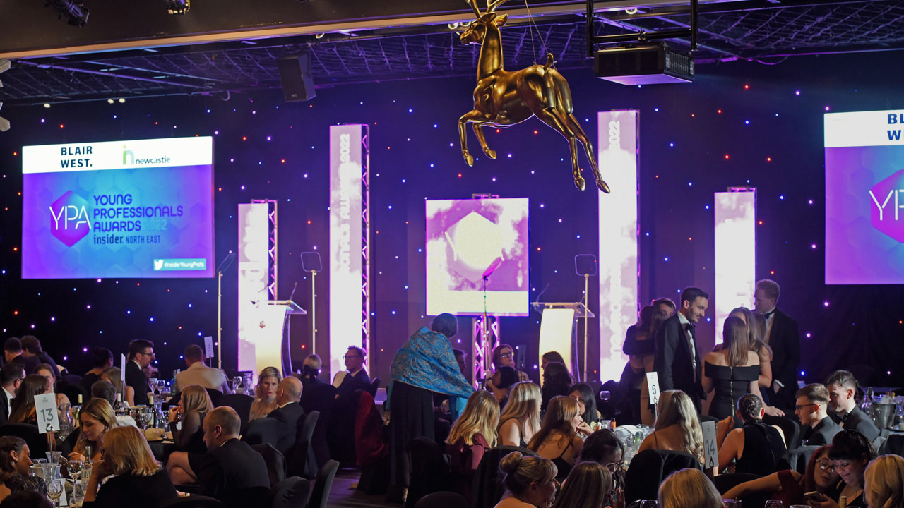 Young Professionals Awards event featuring a decorated venue with stage, purple and blue lighting, golden deer sculpture, and attendees seated at round tables. Two screens display "Blair West" and "Young Professionals Awards" with branding logos. Networking and formal attire set the elegant atmosphere.