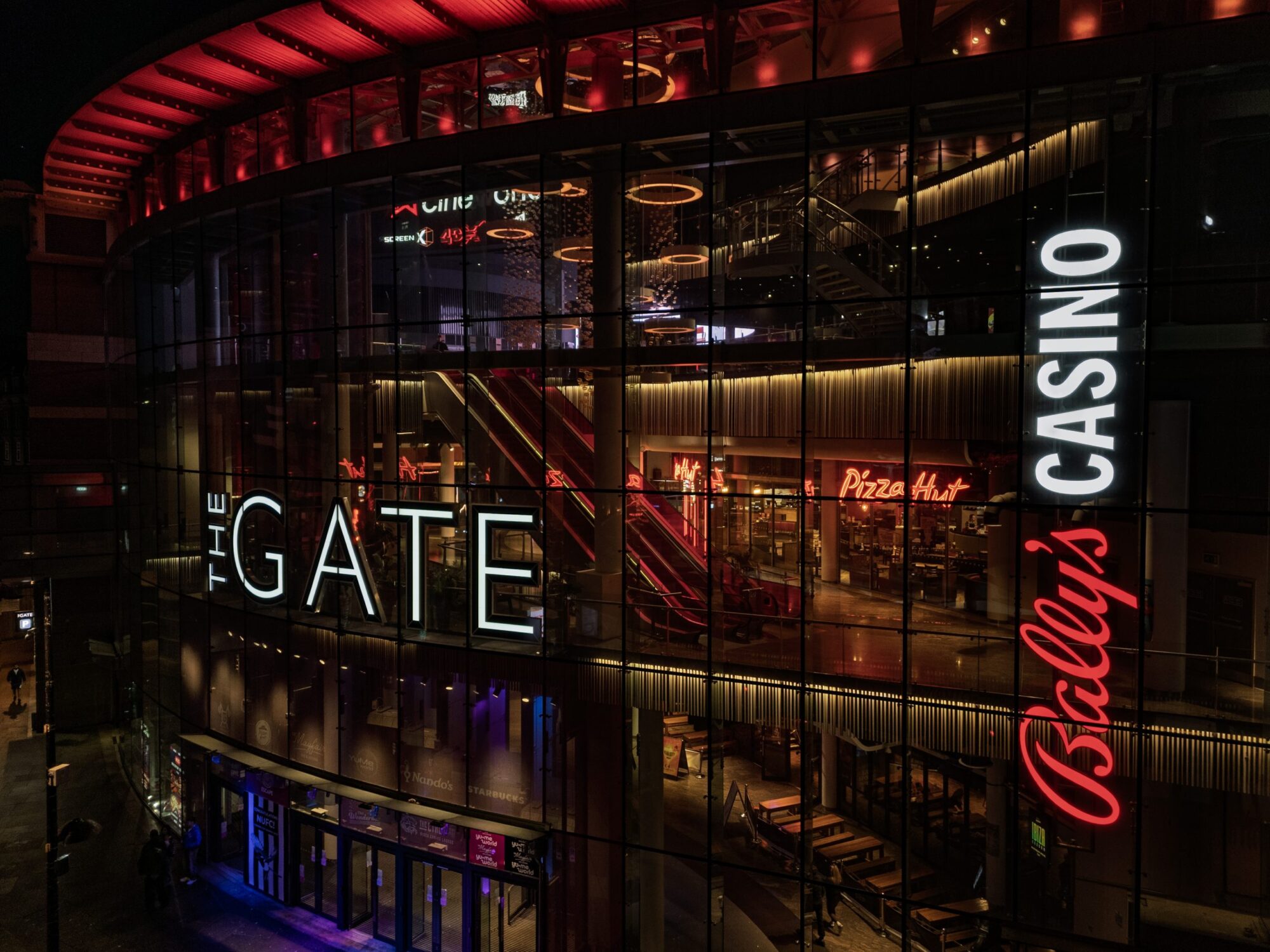 The illuminated facade of The Gate with signs for Pizza Hut and Bally's Casino at night.