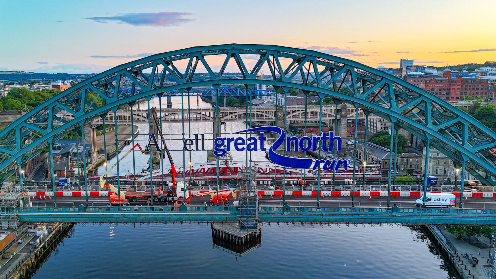 A large arched bridge with "Great North Run" banner spans over a river at sunset.