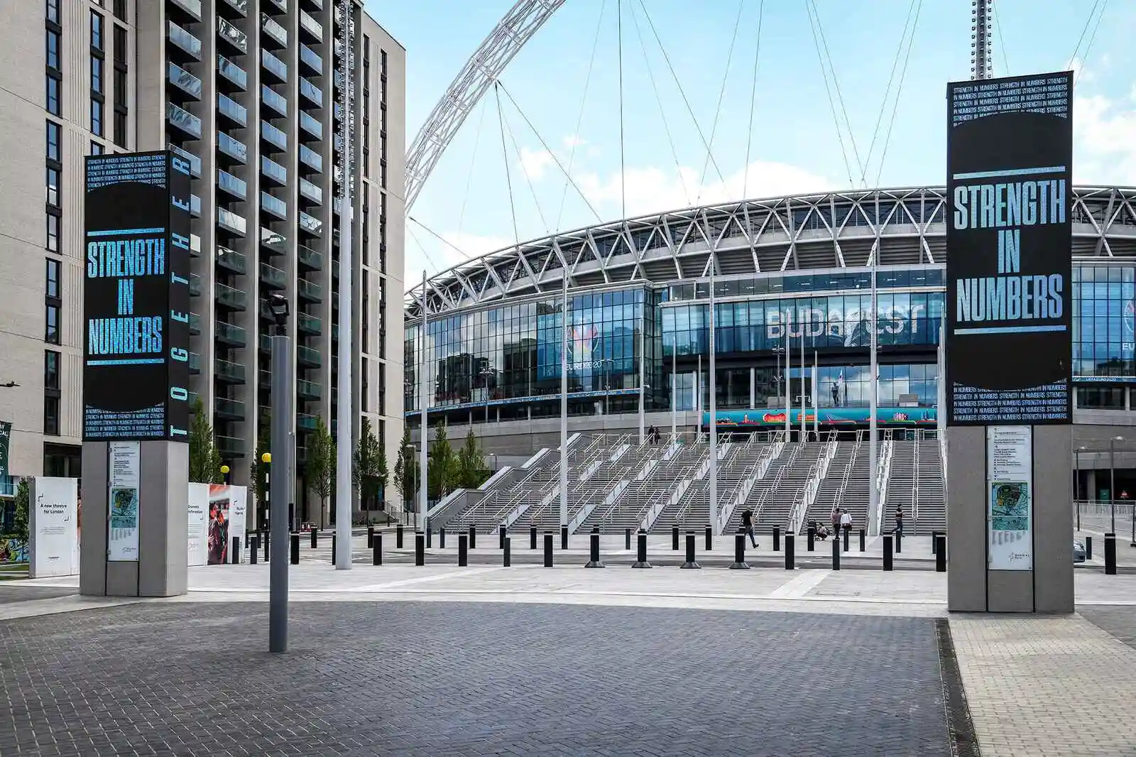 Wembley Stadium entrance with "Strength in Numbers" signs and surrounding modern buildings.