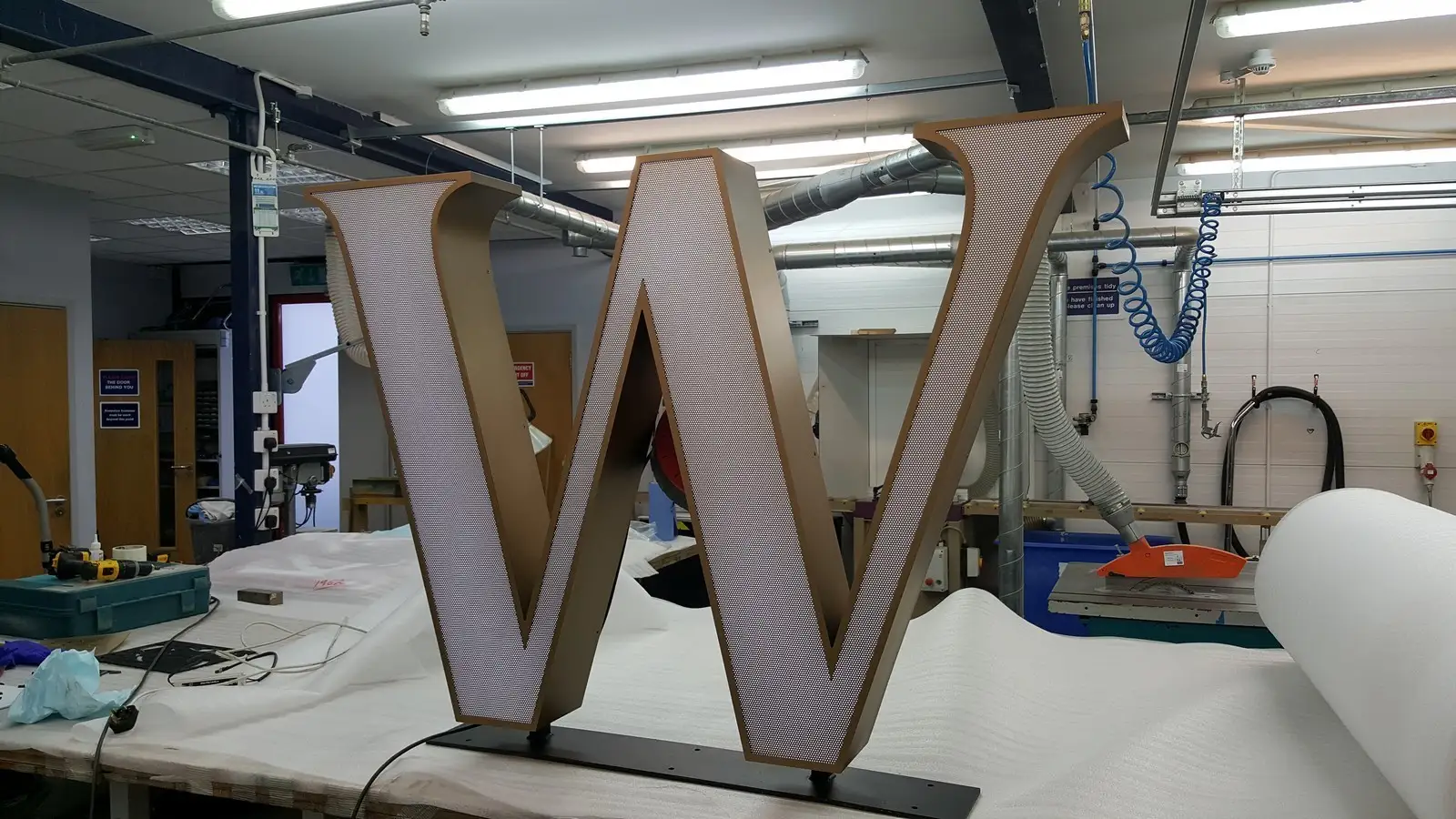 Illuminated large 'W' sign on workbench in industrial workshop with tools and equipment; overhead lighting and surrounding piping visible.