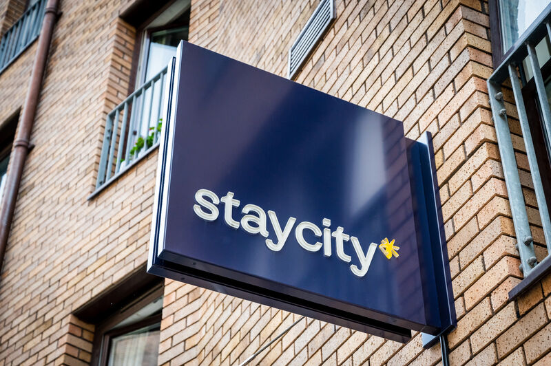 Staycity sign with white text and yellow star logo on a dark blue background, mounted on a brick building facade, next to windows with metal railings and plants.