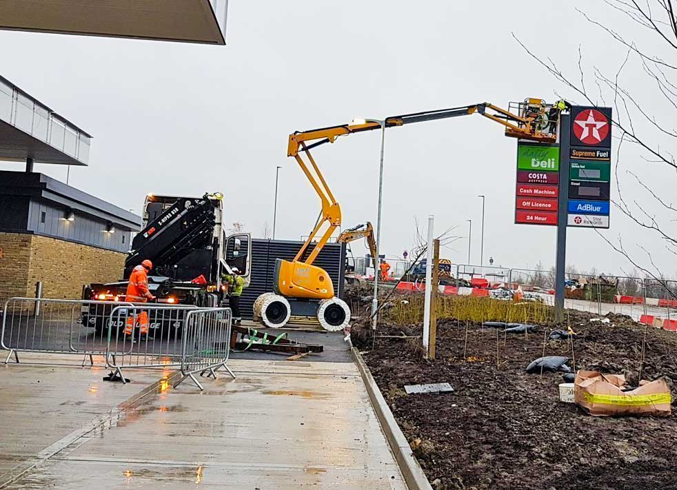 Construction workers in orange suits operate machinery near a Texaco sign on a rainy day.