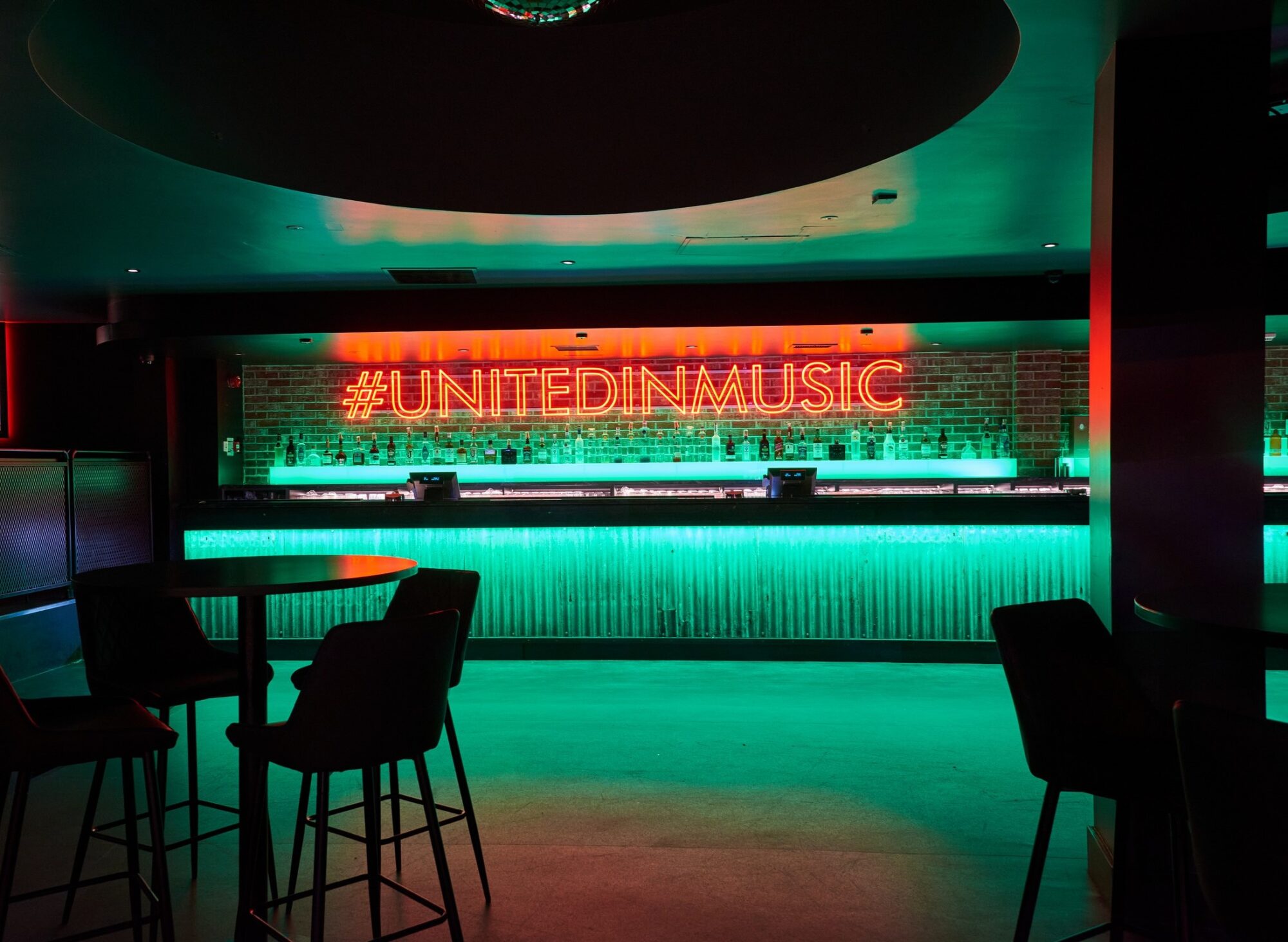 Dimly lit bar with neon sign "#UNITEDINMUSIC" above a counter, surrounded by tables and chairs.