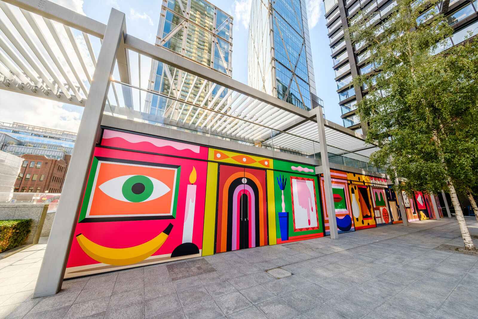 Colorful mural with abstract shapes and patterns on a wall beneath modern glass buildings.