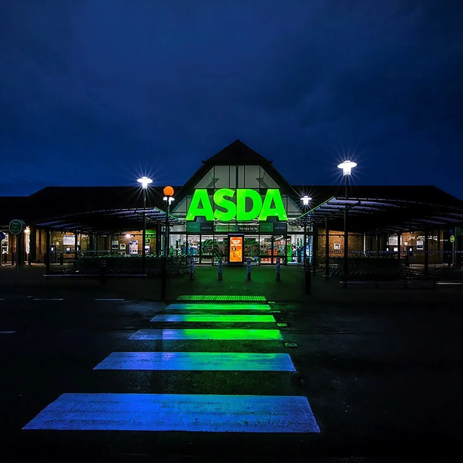 ASDA store entrance illuminated at night with green and white lights, featuring a crosswalk.