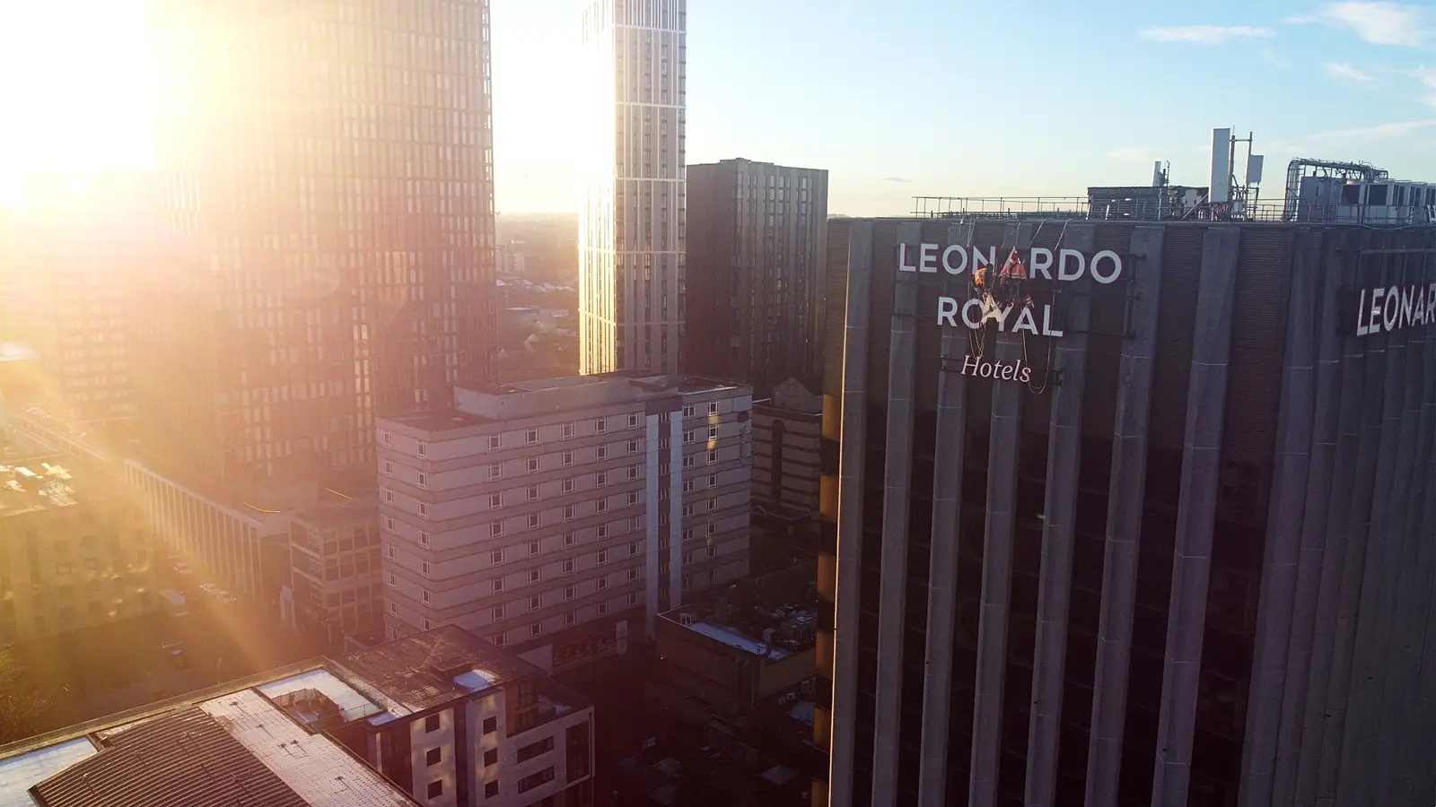 Sunlit cityscape with tall buildings, including Leonardo Royal Hotels, under a clear blue sky.