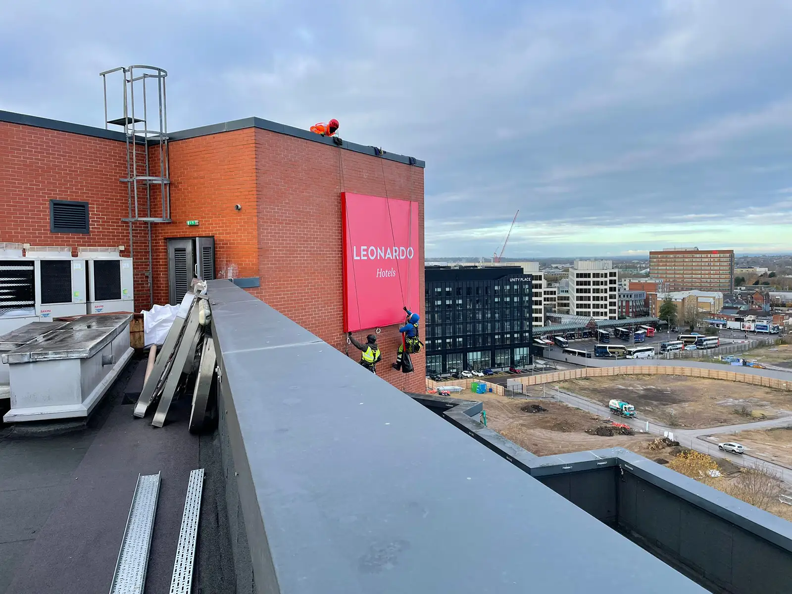 Workers on a rooftop install a large pink sign reading "LEONARDO" on a brick wall.