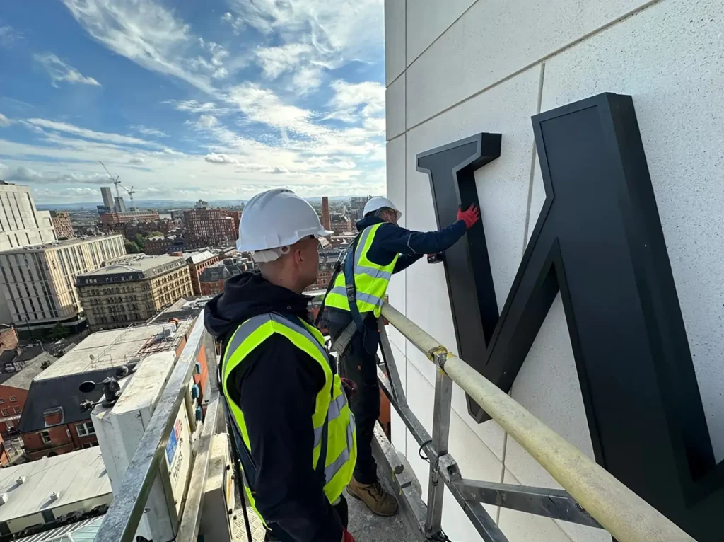 Two workers in safety gear install a large letter "N" on a building facade.