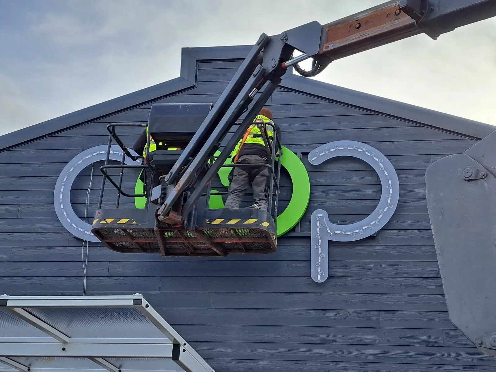 Two workers on a lift install a sign on a dark gray building exterior.