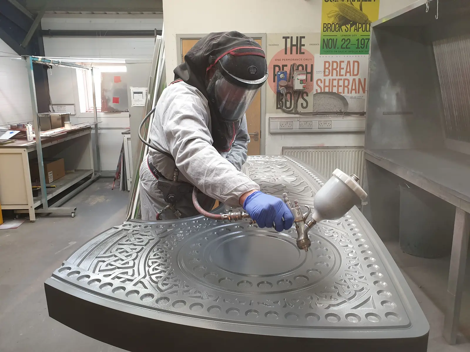 Person in protective gear spray-painting an intricately designed metal surface in a workshop.