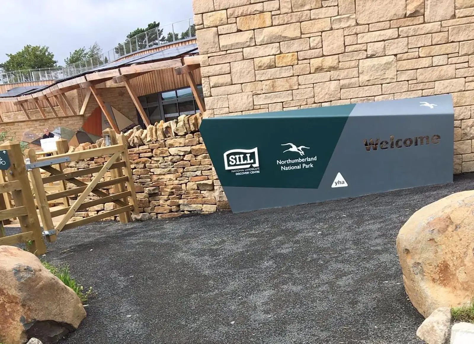 Stone building with a sign reading "Welcome" at Northumberland National Park, near a wooden gate.