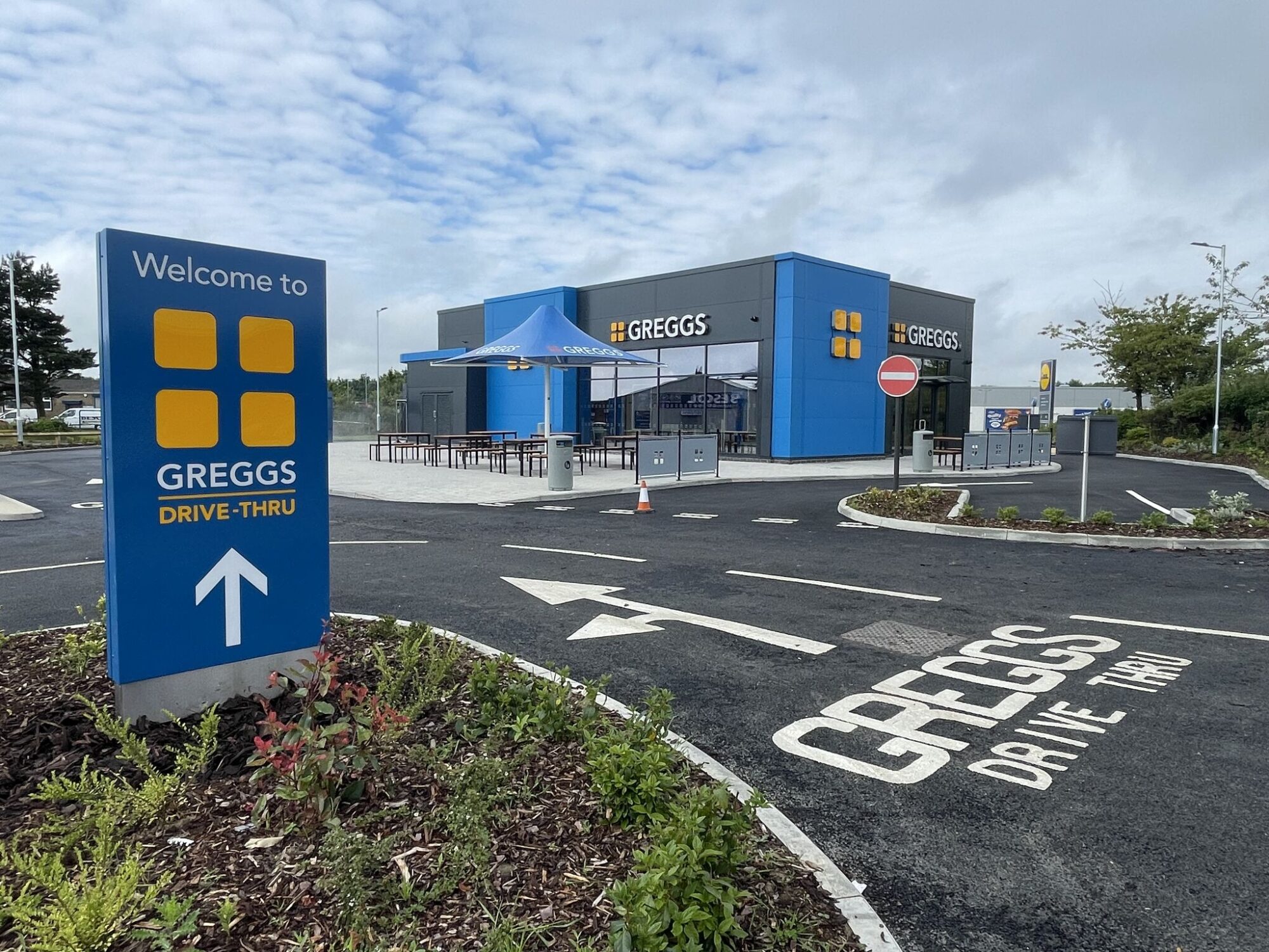 Greggs drive-thru with blue signage, outdoor seating, and directional arrows on the asphalt.