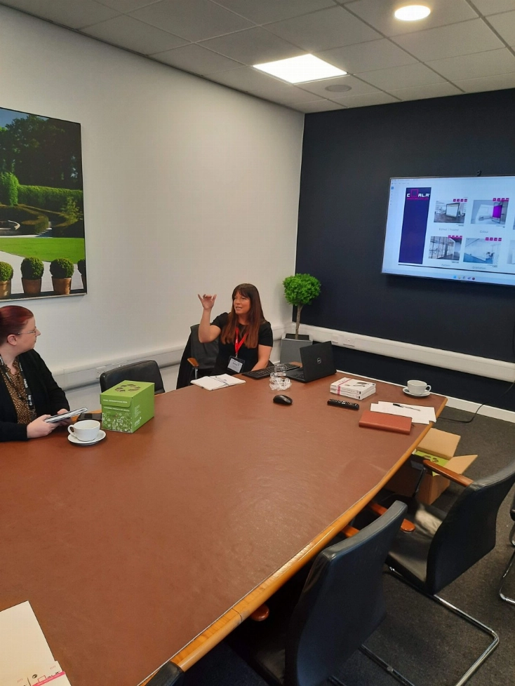 Two people sit at a conference table with a presentation on a wall-mounted screen.