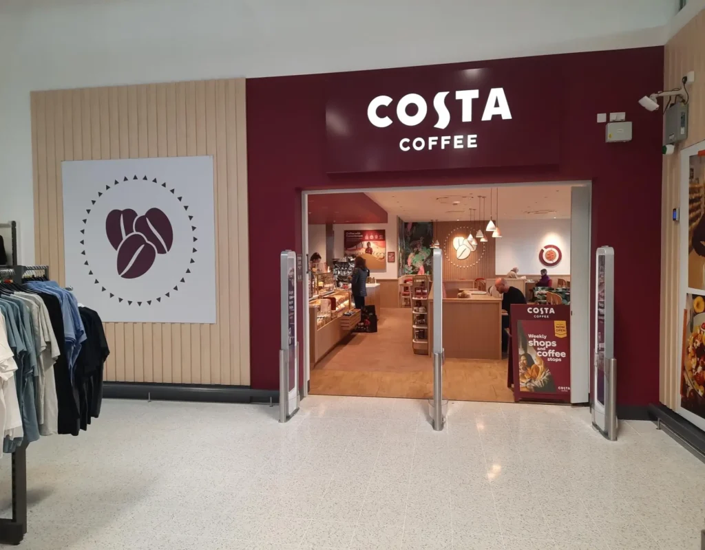 Entrance of a Costa Coffee shop with people inside and clothing racks nearby.