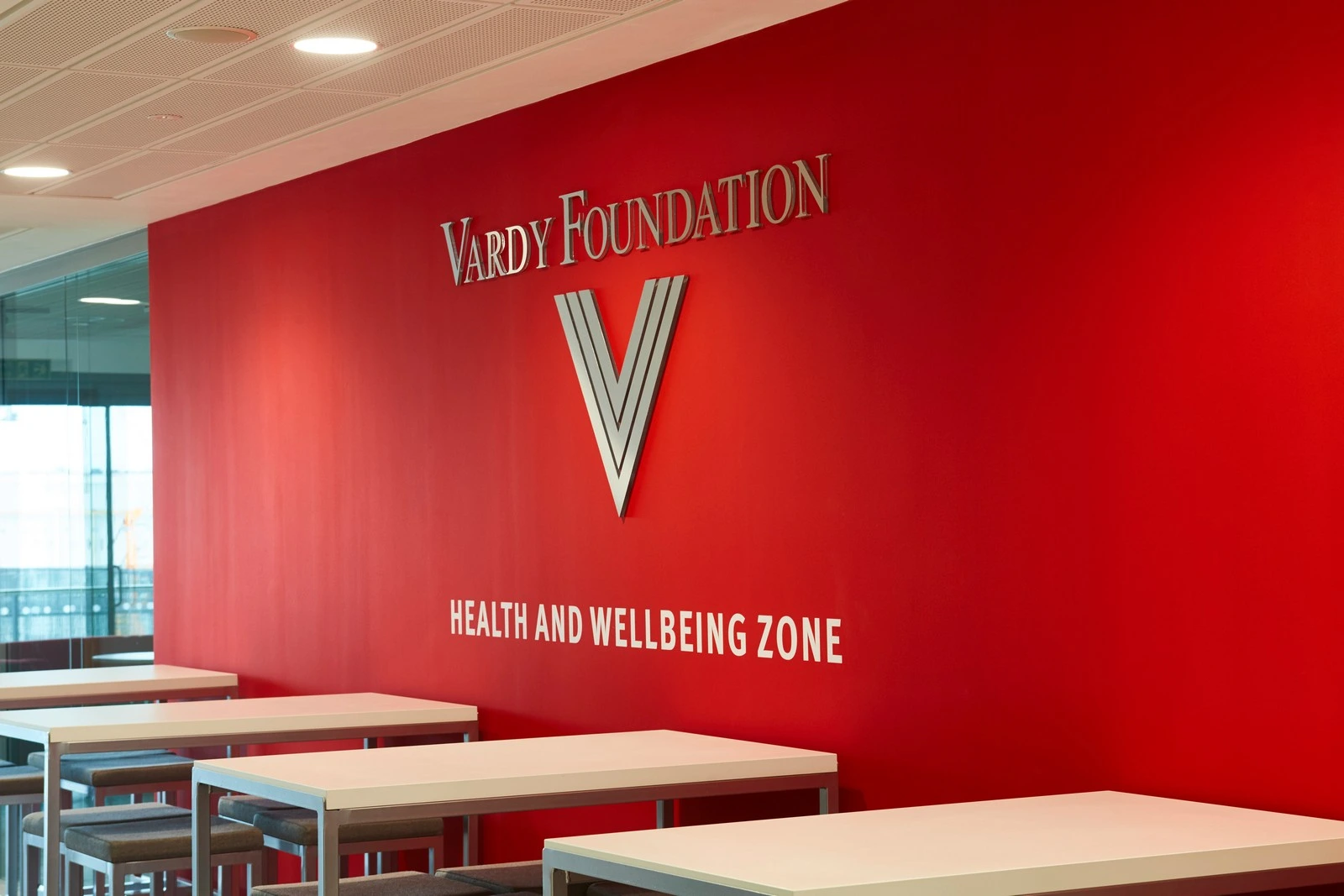 Red wall with "Vardy Foundation Health and Wellbeing Zone" text above white tables.