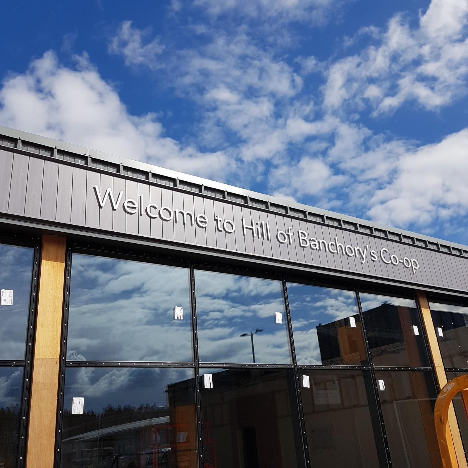 Building facade with "Welcome to Hill of Banchory's Co-op" sign under a blue sky.