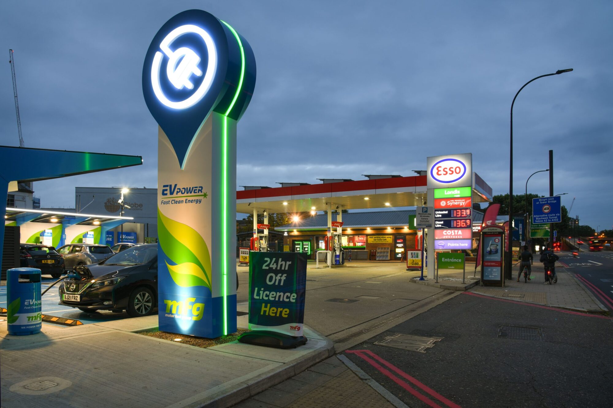 A gas station with EV charging, illuminated signs, and a convenience store at dusk.