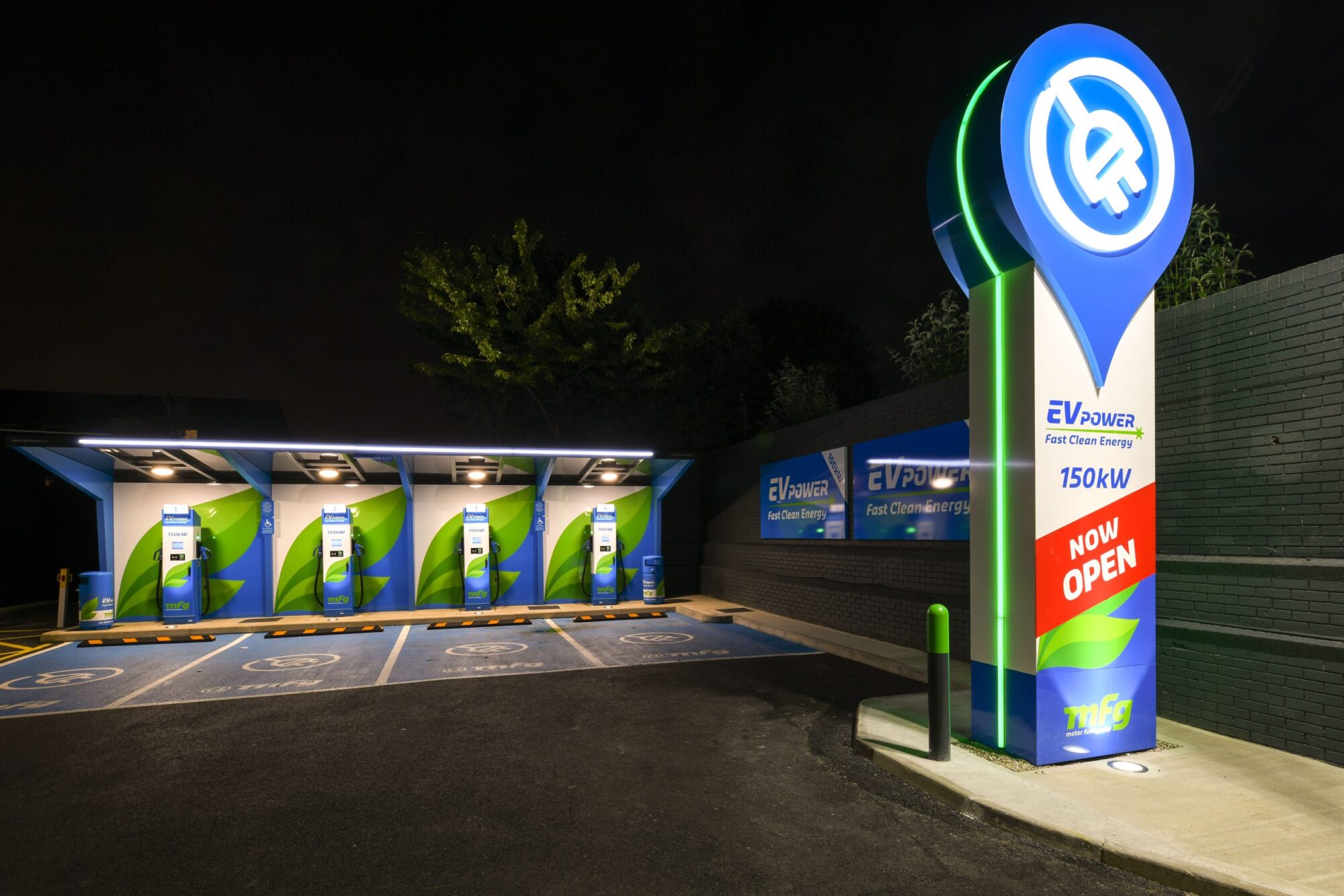 Electric vehicle charging station at night with multiple charging units and a "Now Open" sign.