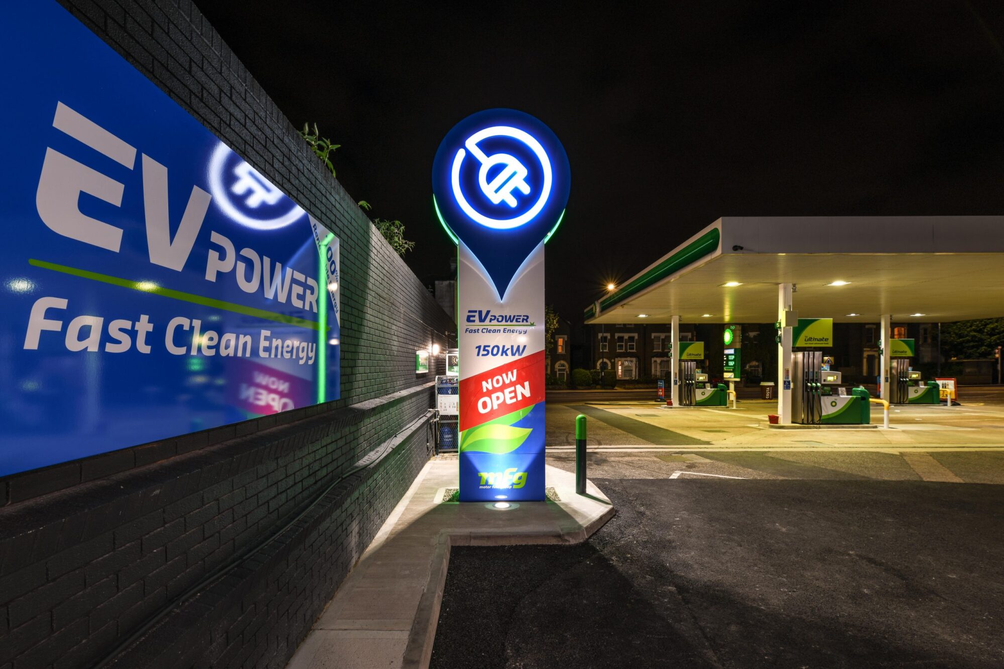 Electric vehicle charging station with illuminated signs and a gas station in the background at night.