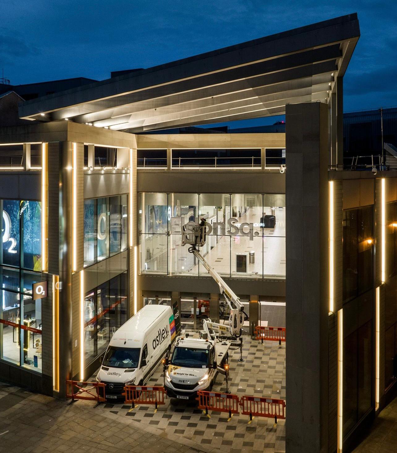 A modern building with large windows is illuminated at night, featuring two parked service vehicles.