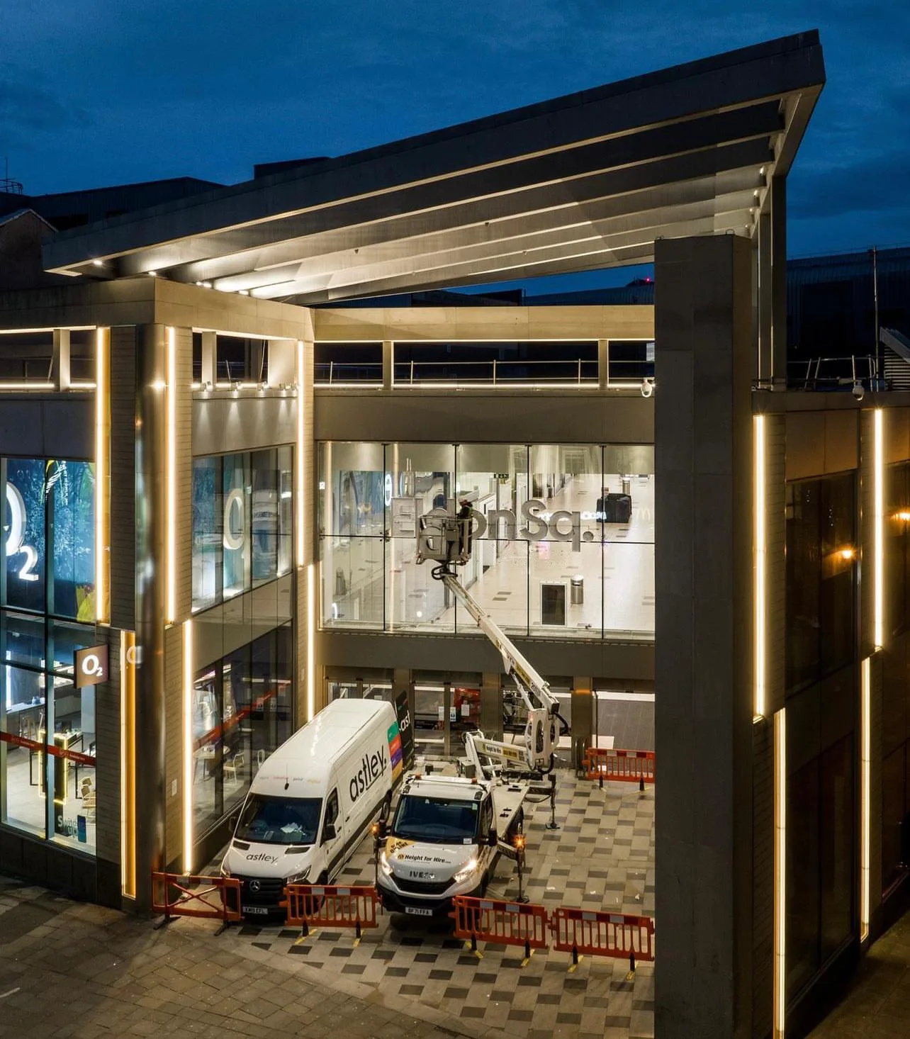 A modern building with large windows is illuminated at night, featuring two parked service vehicles.