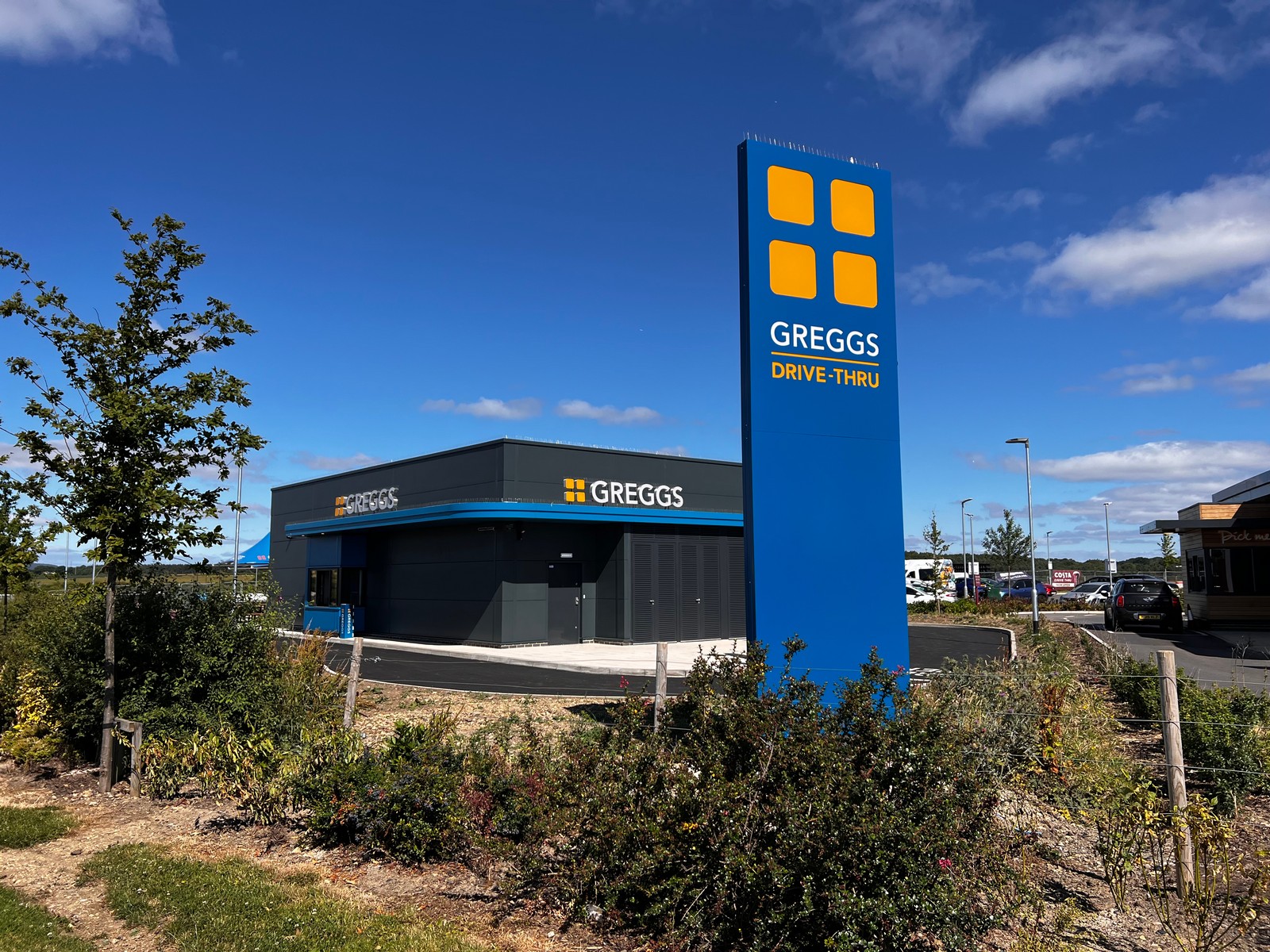 A Greggs drive-thru with a tall blue sign and surrounding greenery under a blue sky.