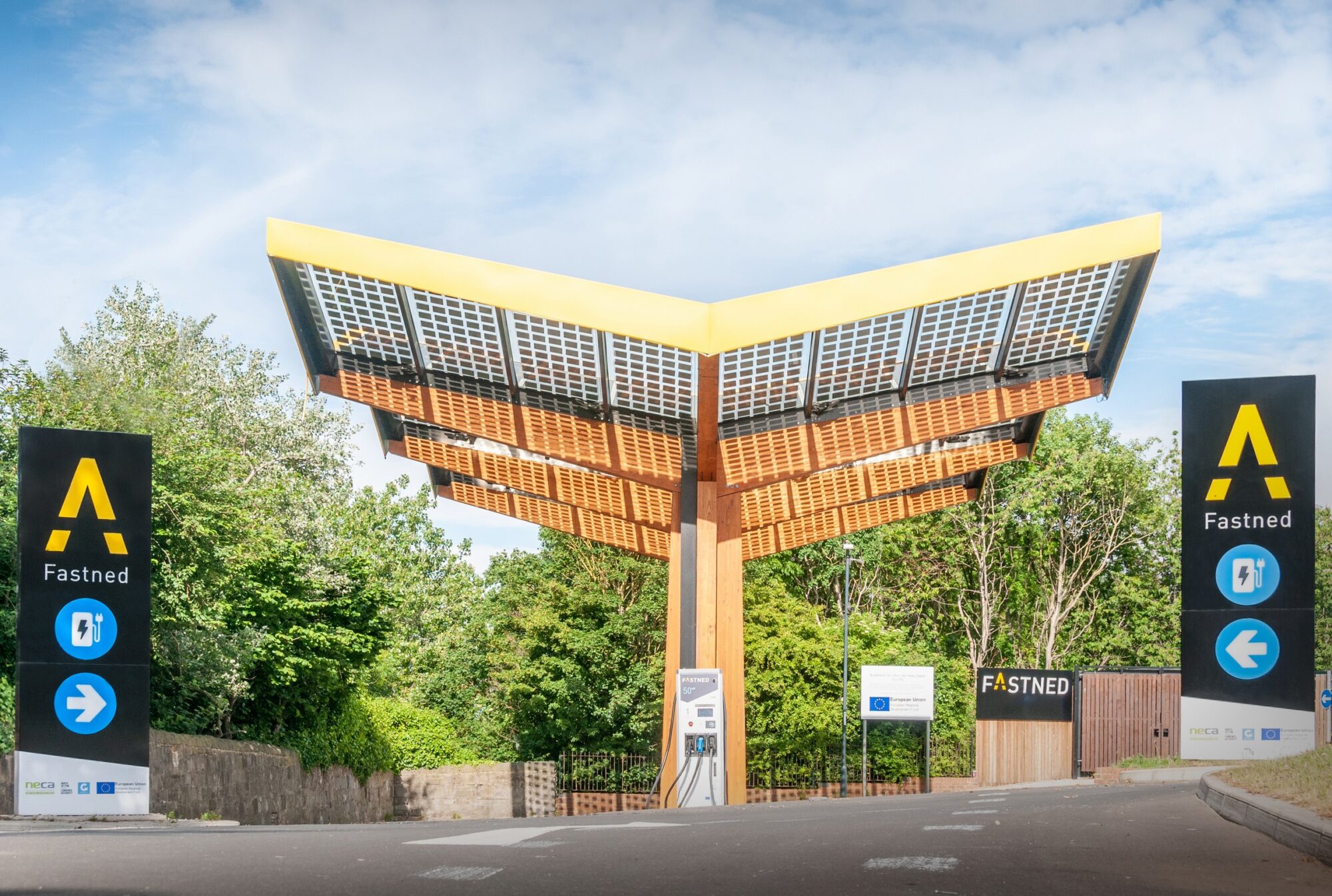 A solar-powered Fastned charging station with a distinctive V-shaped roof amidst greenery.