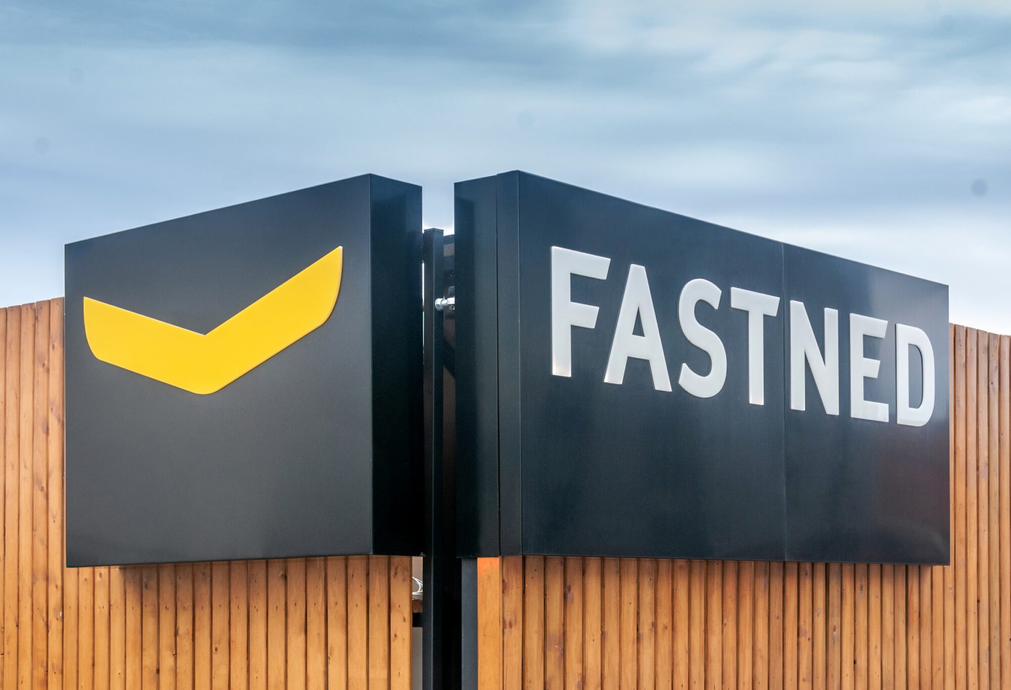 A black sign with "FASTNED" and a yellow logo stands against a wooden fence and cloudy sky.