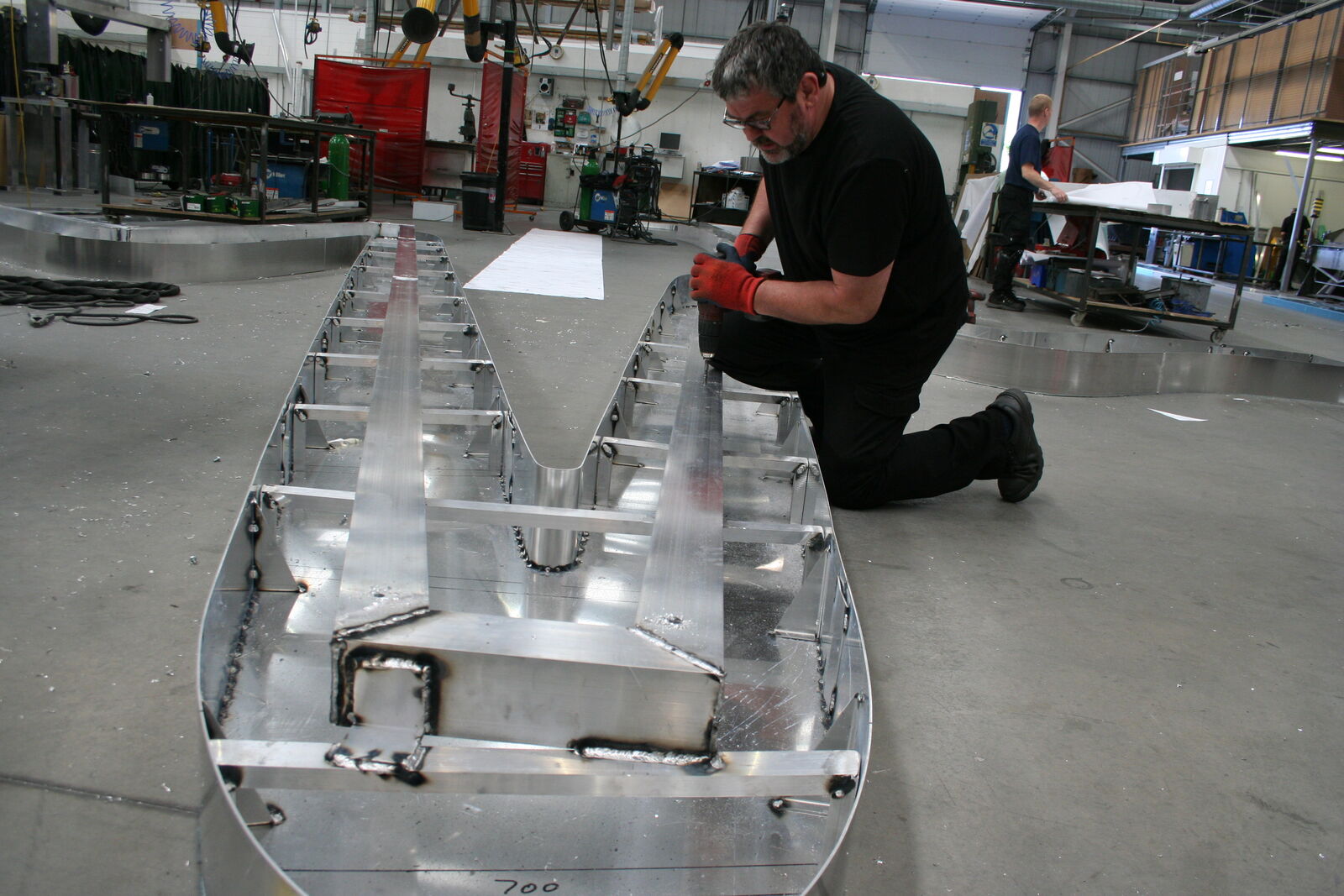 A man kneels beside a large metal structure in an industrial workshop.