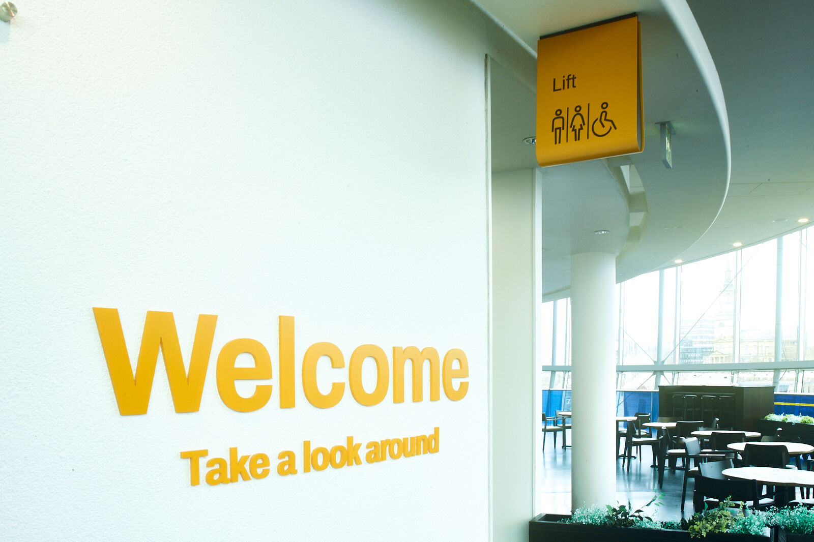 Yellow "Welcome" sign on a white wall near a lift sign and seating area.