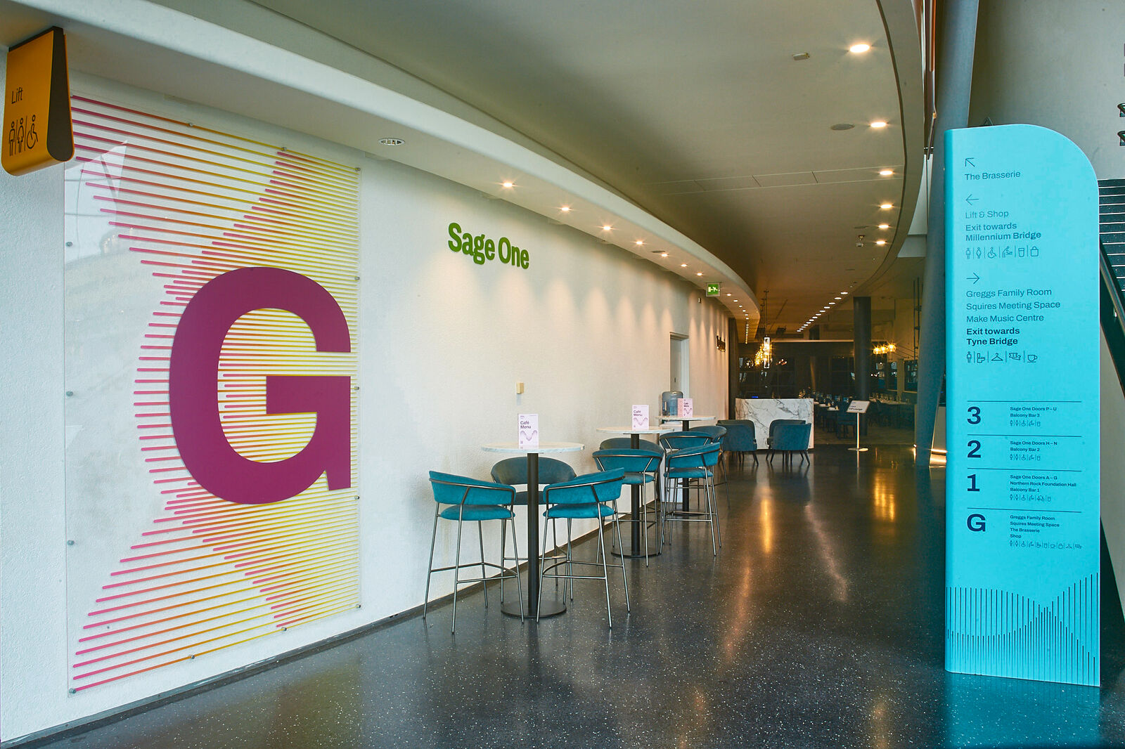 A hallway with "Sage One" sign, tables, chairs, and a large "G" on the wall.