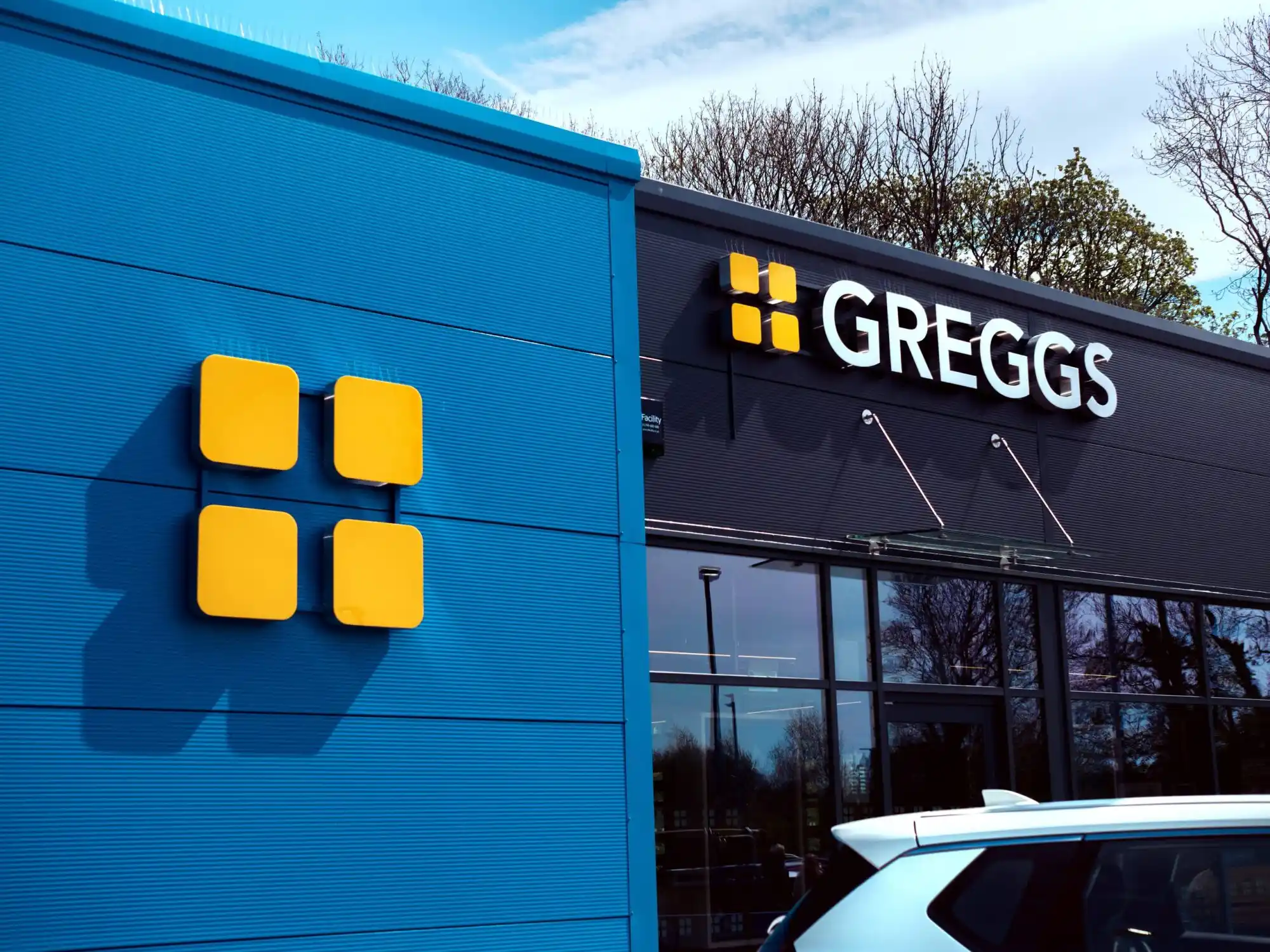 Blue and black building with yellow square logo and "Greggs" sign, trees in background.