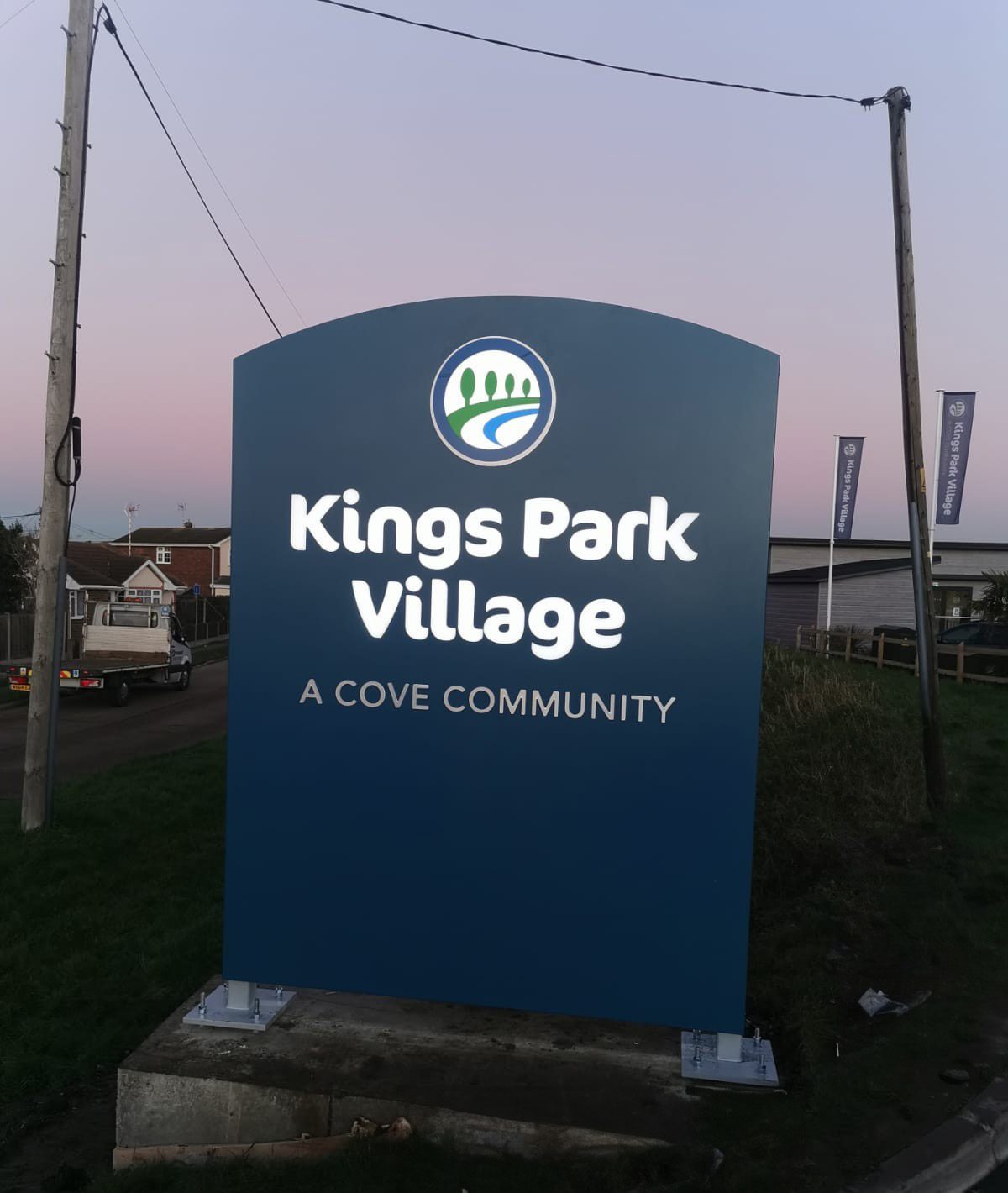 Blue sign reading "Kings Park Village, A Cove Community" with a logo, near a road.