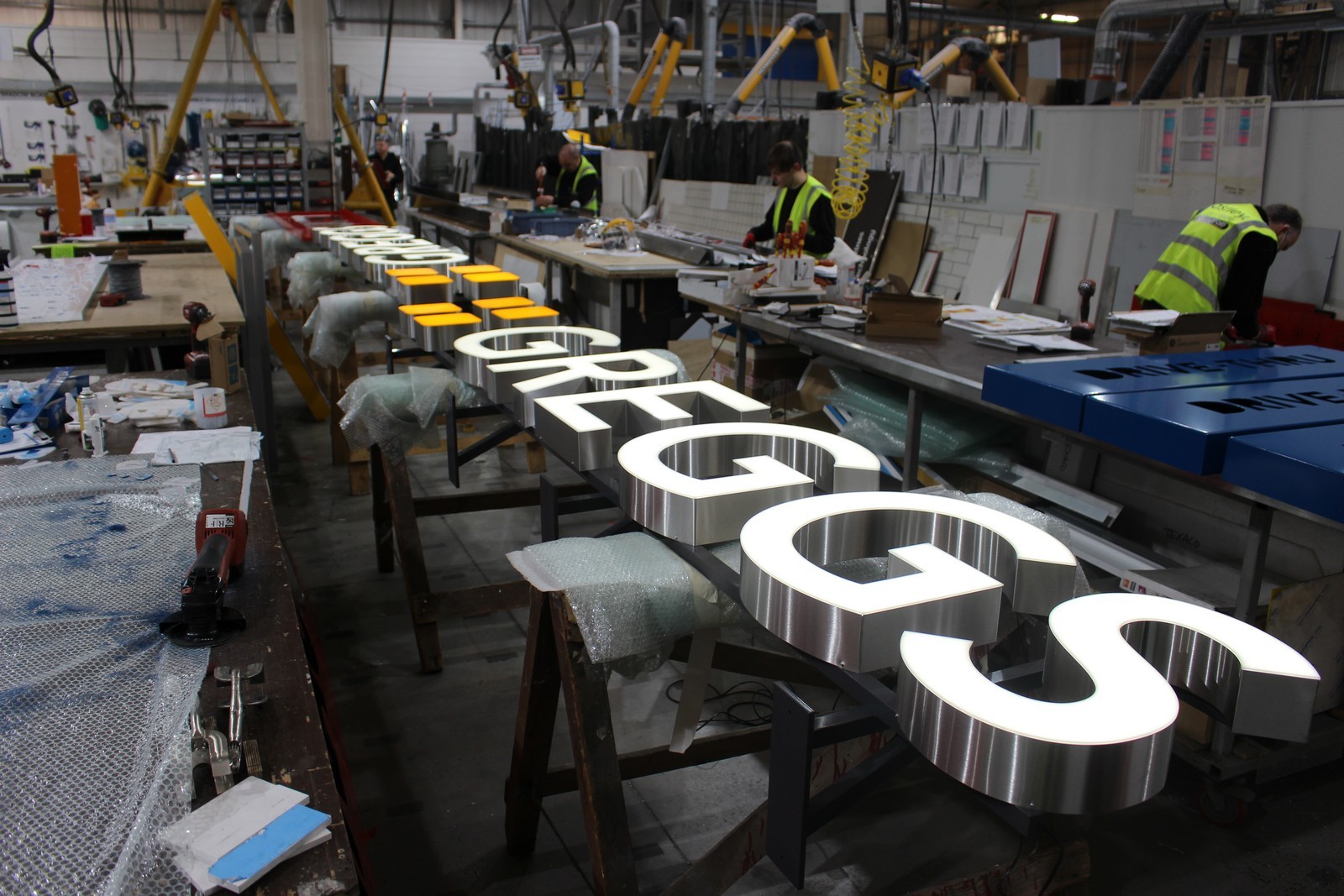 Workers in a factory assemble large illuminated letters spelling "GREGGS" on a long table.