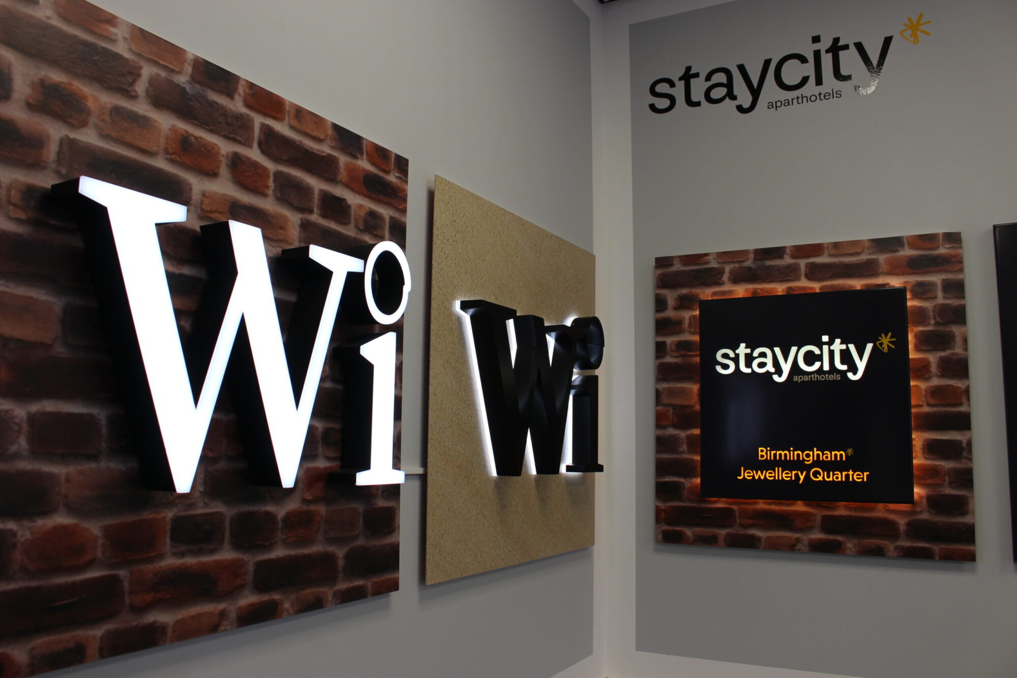 Illuminated "Wi-Fi" sign in front of brick-patterned paneling, adjacent to a "staycity aparthotels" sign. The staycity sign is accompanied by text "Birmingham Jewellery Quarter" beneath, suggesting location branding.