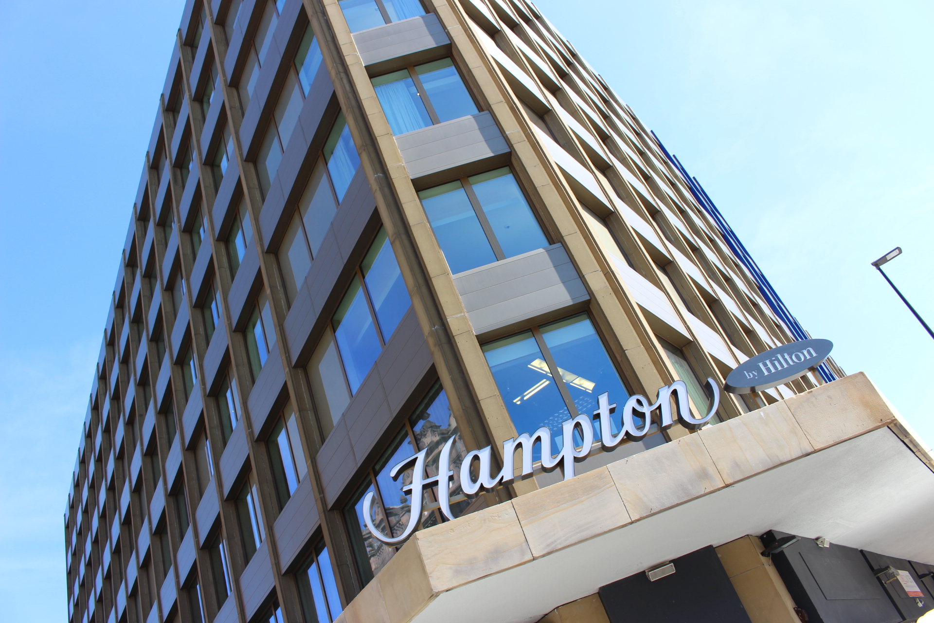 Tall building with "Hampton by Hilton" sign, featuring numerous windows against a clear blue sky.