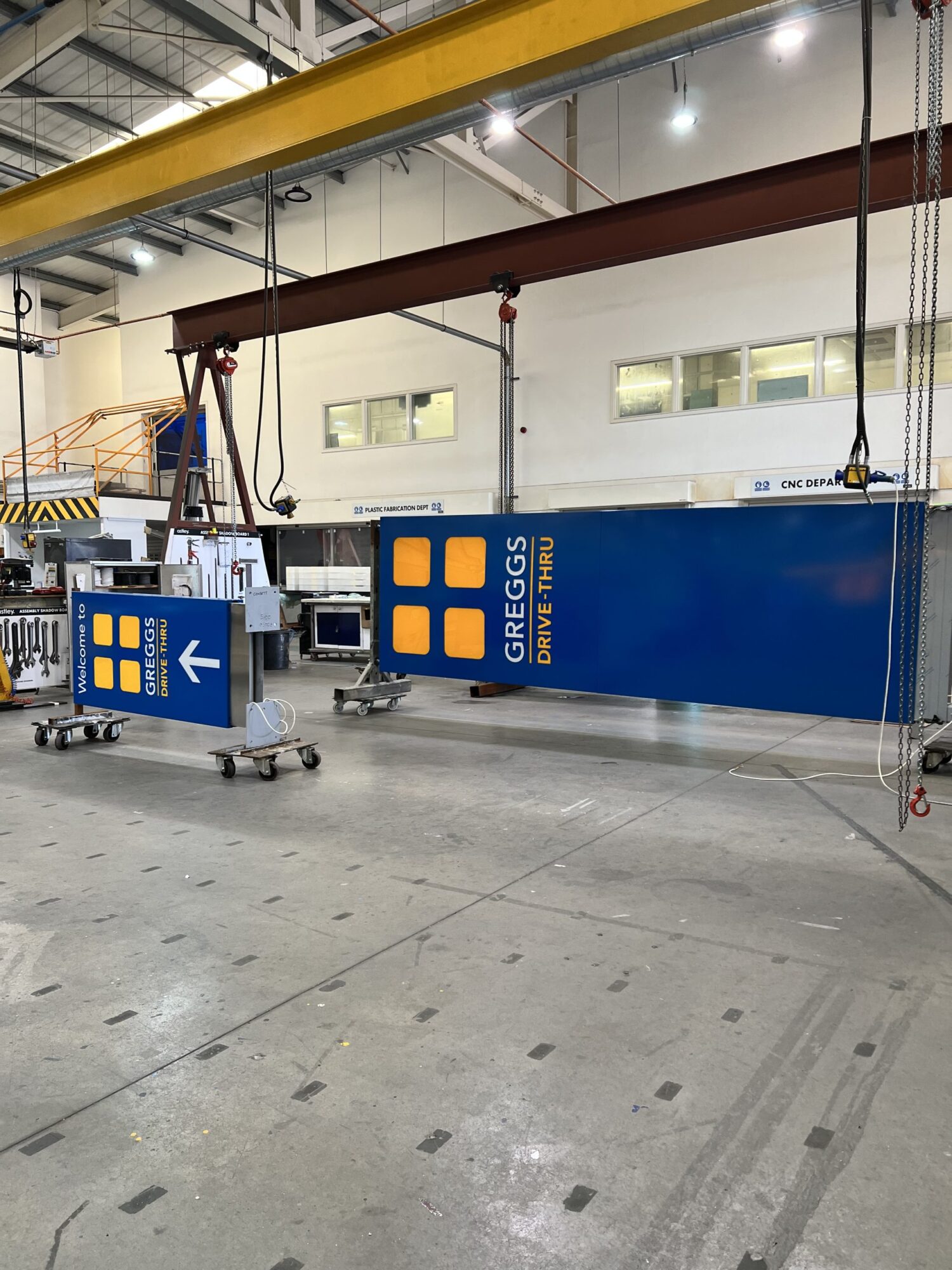 Large blue "Greggs Drive Thru" sign suspended in an industrial warehouse setting.