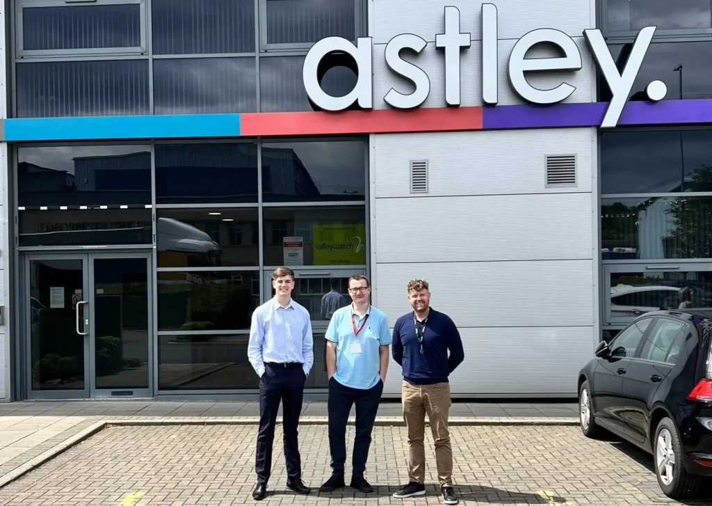 Three men standing in front of a building with the sign "astley.