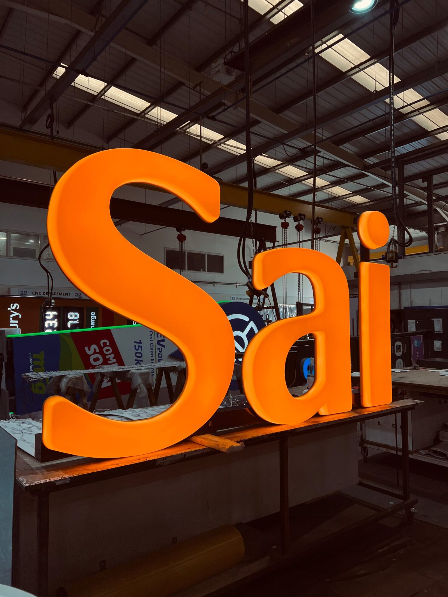 Large illuminated orange letters spelling "Sai" in an industrial workshop setting.