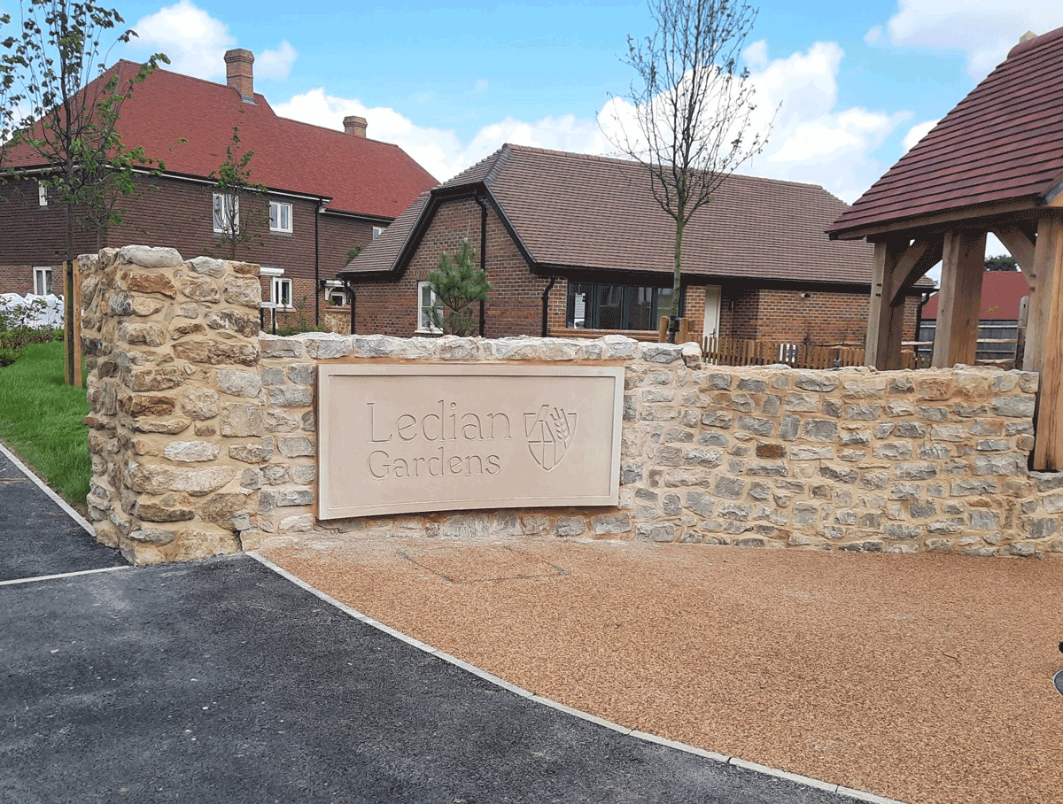 Stone wall with "Ledian Gardens" sign, surrounded by houses and trees under a blue sky.
