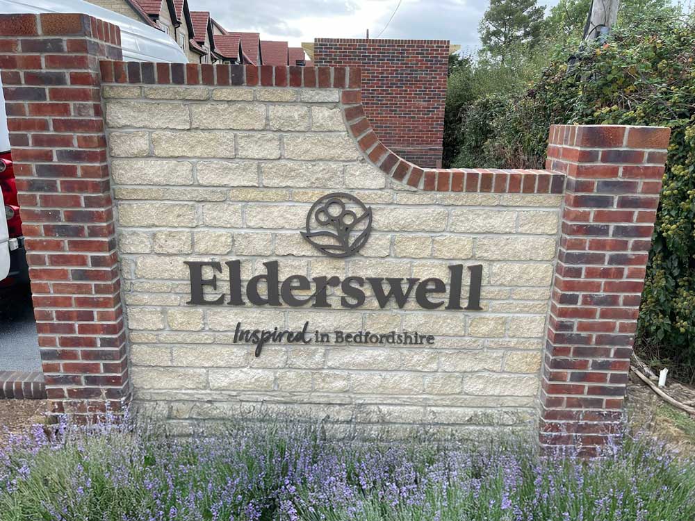 Stone sign with "Elderswell inspired in Bedfordshire" surrounded by brick pillars and lavender plants.