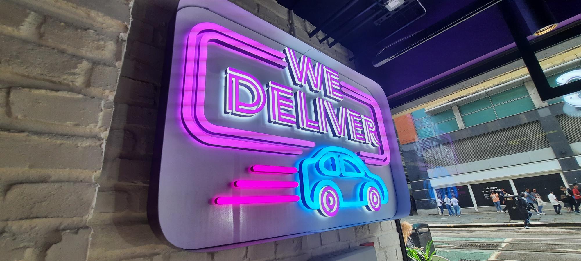 Neon sign reading "We Deliver" with a car graphic, mounted on a brick wall.