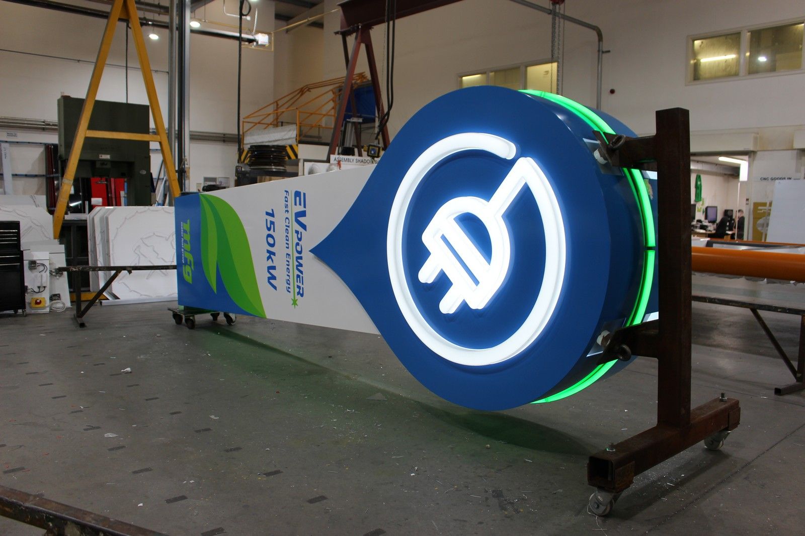 Large EV charging station sign with a neon plug icon, lying on a workshop floor.