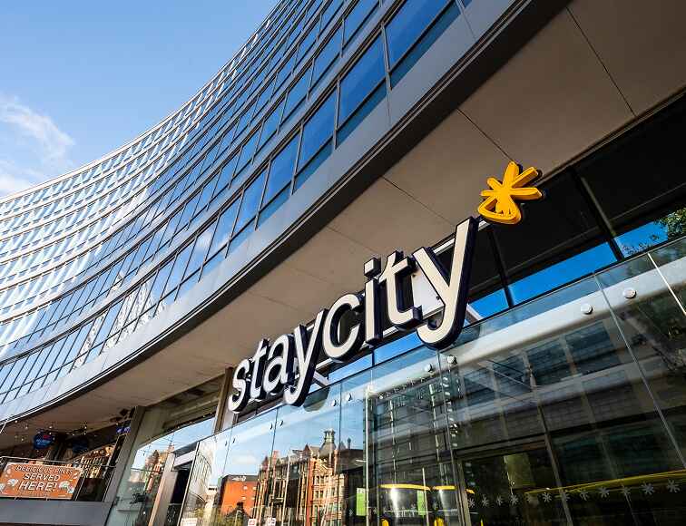 Curved modern building with large "staycity" sign and reflections of surrounding architecture in glass.