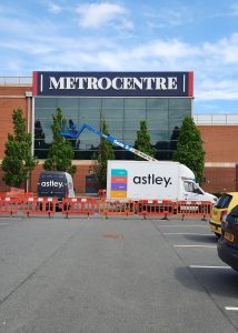 Workers on a blue lift are installing a sign on the Metrocentre building facade.